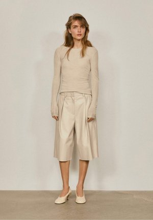 Femme debout contre un mur uni, portant un haut beige à manches longues, un short beige large jusqu'aux genoux et des chaussures à talons beige.