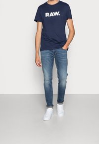 Navy blue cotton T-shirt with "RAW." in large white letters, paired with light blue jeans and white sneakers, standing on a neutral floor.