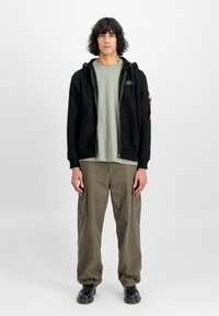 Sort zip-up hoodie med frontlommer, båret over en lys grøn T-shirt og parret med olivengrønne cargo-bukser. Sorte sko fuldender outfittet.