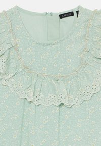 Mintgrønn bluse med blomstermønster, med rynkete utsnitt ved halsen og blonder. Lett stoff med teksturerte mønstre. Glidelås i ryggen.
