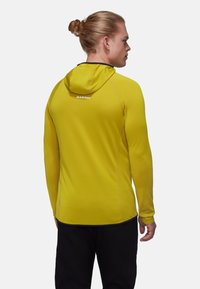 Ljusgul huvtröja i texturerad tyg, med platta sömmar och kontrasterande svart kant runt ärmsluten och nederkanten. "Mammut"-logotyp på ryggen.