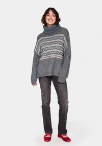 Pulover din tricot în tonuri de albastru cu guler tip cowl, având un model geometric în gri deschis și teal. Asortat cu blugi închisi la culoare și balerini roșii.