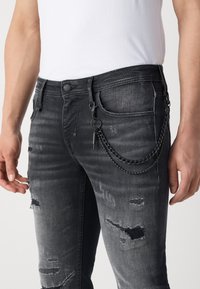 Homme portant un jean noir usé avec une chaîne noire attachée à la poche avant et à la boucle de ceinture, associé à une chemise blanche unie rentrée.