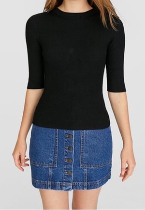 Vrouw draagt een aansluitend zwart geribbeld topje met driekwart mouwen en een blauwe denim minirok met knopen aan de voorkant en grote voorzakken.