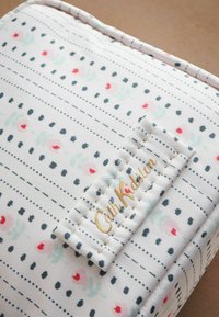 Katoenen tas met een witte achtergrond, versierd met meerkleurige bloempatronen en stippen, en een gouden logo-tag. Geweven, getextureerd ontwerp.