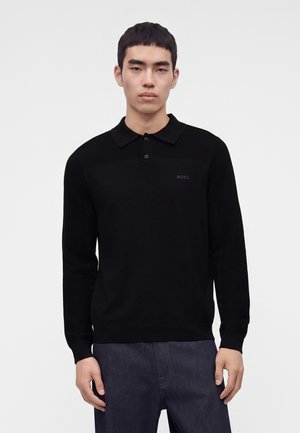 Junger Mann mit kurzen schwarzen Haaren, trägt ein schwarzes langärmeliges Poloshirt und dunkle Hose, steht vor einem hellen, einfarbigen Hintergrund.