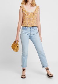 Gul blus med blommönster och volangkrage, i kombination med ljusblå raka jeans och svarta sandaler med remmar. Håller en liten vävd handväska.