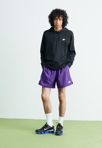 Mladenič oblečen v črn Nike kapuco, vijolične Nike kratke hlače, bele nogavice in črne Nike superge stoji na zeleni podlagi pred belo ozadjem.