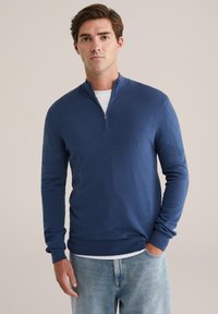 Pulover cu guler tip fermoar de culoare bleumarin, realizat dintr-un material moale și texturat, având mâneci lungi și tiv zimțat, asortat cu blugi decolorati.