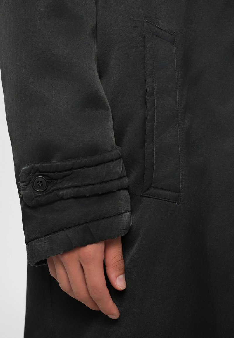 Cappotto nero con una texture liscia, dotato di polsini a risvolto e un dettaglio con bottone. C'è una tasca laterale visibile sul corpo.