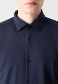 Camisa de botones azul marino con cuello extendido, textura suave y botones negros en la tapeta frontal, con un corte ajustado.