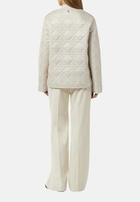 Helle beige gesteppte Jacke mit strukturierter Oberfläche, rundem Ausschnitt und Logodetail auf dem Rücken. Kombiniert mit weit geschnittenen cremefarbenen Hosen.