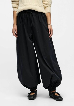 Personne portant un pantalon large noir à jambes ballons avec des poignets élastiques, un pull beige, des ballerines noires Mary Jane et un bracelet en argent.