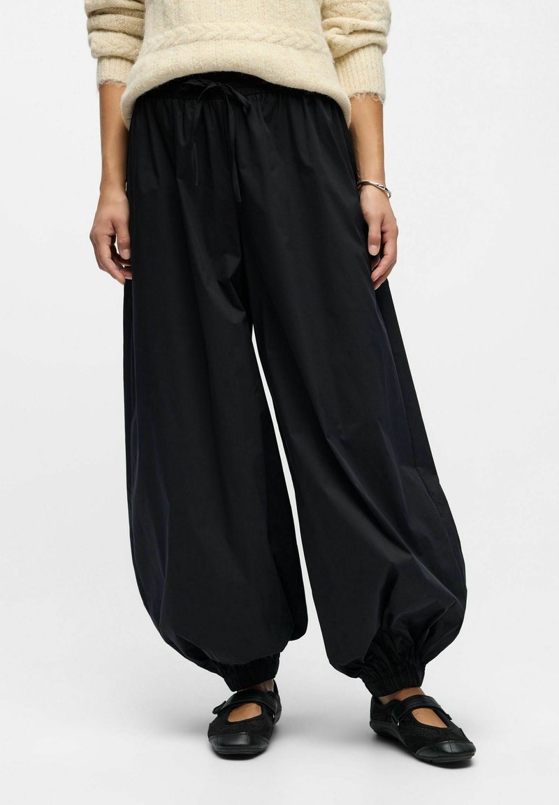 Person, der lockere schwarze Ballonhosen mit elastischen Bündchen, einen beigen Pullover, schwarze Mary Jane Ballerinas und ein silbernes Armband trägt.
