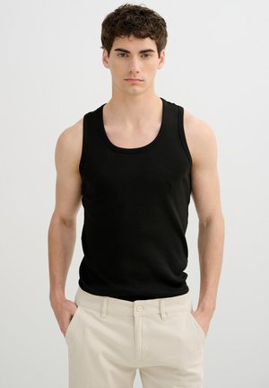 Joven con cabello corto y rizado, vestido con una camiseta sin mangas negra y pantalones beige claros, de pie frente a un fondo claro y liso.