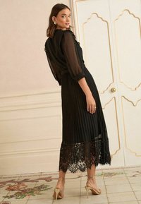 Robe noire plissée avec un ourlet en dentelle, des manches longues en voile et uncol en volants ; accompagnée de talons dorés à lanières. Conception élégante et féminine.