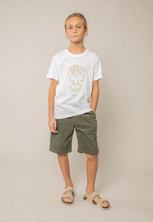Jeune garçon debout avec une main dans la poche, portant un t-shirt blanc avec un motif de crâne fleuri, un short vert et des sandales beige sur un fond neutre.