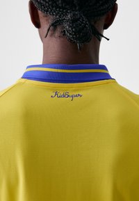 Maglia gialla con tessuto testurizzato. Colletto e rifiniture blu con due righe gialle. Logo "KidSuper" ricamato in modo discreto in blu.