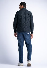 Schwarze gesteppte Jacke mit hohem Kragen, langen Ärmeln und strukturiertem Muster, kombiniert mit blauen Denim-Hosen und beigen Schuhen.