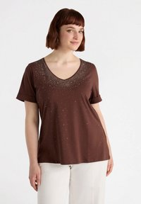 Camiseta corta de manga corta color marrón con escote en V, que presenta adornos plateados dispersos cerca del escote y un tejido suave y texturizado.