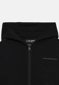 Sweatshirt preto com fecho, de textura suave, com capuz, fecho à frente e um pequeno logótipo no peito. Feito de tecido macio.