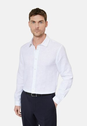 Mężczyzna ubrany w białą, długorękawową koszulę zapinaną na guziki wpuszczoną w czarne spodnie z czarnym paskiem, stoi z jedną ręką w kieszeni.