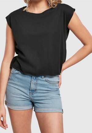 Frau trägt ein schwarzes ärmelloses Top und hellblaue hochgekrempelte Jeansshorts, steht mit einer Hand in der Hüfte.