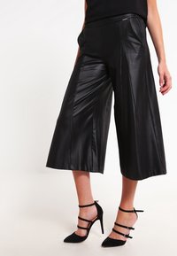 Culottes en simili cuir noir avec une coupe à jambes larges, présentant des plis avant et des poches latérales. Associées à des chaussures à talons noires.