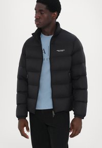 Black puffer jacket with a high collar, horizontal quilted design, logo on the chest, and zippered pockets. Worn over a light blue shirt.