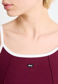 Bordeļa tank tops ar baltām siksnām, iekļaujot teksturētu adījuma rakstu un melnu "obey" uzlīmi priekšā.