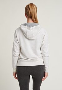 Felpa con cappuccio grigia chiara con cappuccio interno grigio a contrasto. Presenta maniche raglan e una texture liscia. Abbinata a leggins scuri.