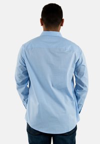 Camisa de botón de color azul claro con cuello, mangas largas y dobladillo curvado. Fabricada de material de algodón texturizado. Se muestra la vista trasera.