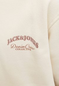 Tessuto color crema con il testo marrone "JACK & JONES Denim Crew COLLECTIVE" stampato sopra, che mostra parte di una cucitura della manica.