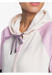 Sudadera con capucha y cremallera en color crema con mangas rosa claro, que presenta un cordón morado y un logo bordado en el lado izquierdo.