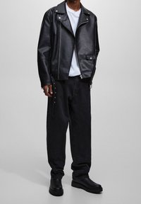 Chaqueta de motorista de cuero negro con herrajes plateados, que presenta un cuello con muesca y bolsillos con cremallera, combinada con vaqueros negros holgados y botas negras.