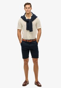 Strik t-shirt med korte ærmer i creme, navy sweater bundet om halsen, navy shorts, brun læderbælte og brune loafers. Afslappet sommeroutfit.