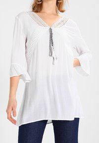 Top bianco fluido con scollatura a V e dettagli in crochet. Presenta un laccio con accenti in bianco e nero. Maniche a tre quarti.