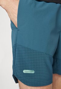 Herrshorts i teal, med ett slätt övre tyg och ett texturerat rutmönster på den nedre delen. "VENTUS"-etikett syns på fållen.
