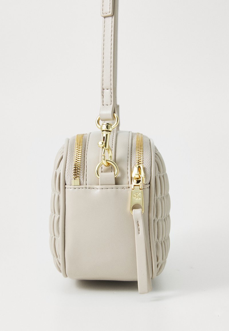 Beżowa skórzana torebka z pikowanym wzorem, złotymi zamkami błyskawicznymi i metalowymi elementami. Zawiera odpinany pasek oraz płaski, prostokątny kształt.