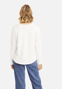 Weißes Langarmhemd mit abgerundetem Saum, aus weichem Stoff, kombiniert mit lockeren blauen Denim-Hosen. Von hinten sind die Raglanärmel sichtbar.