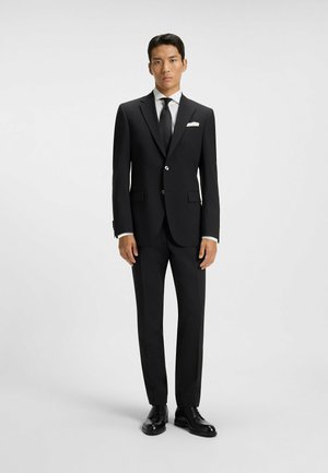 Svart skreddersydd dress med enkel knappelukking, hvit skjorte, svart slips, hvit lommefyr, og svarte lær sko.