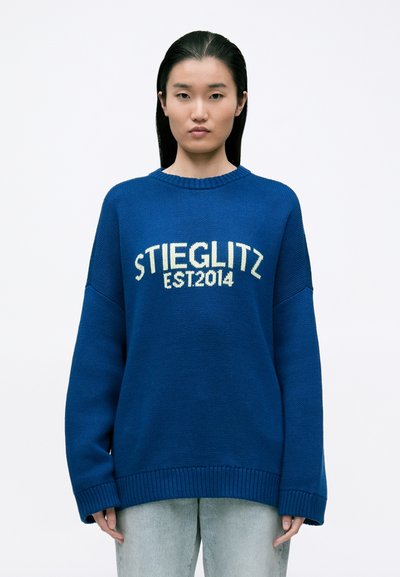 Blå strikket sweater med ribbet hals og kant, der har hvid tekst "STIEGLITZ EST.2014" centralt placeret. Modellen har lyse jeans på.