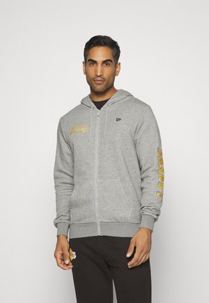 Sudadera gris con cremallera y el texto dorado "Lakers", con bolsillos frontales y capucha con cordón. Se lleva con pantalones negros que presentan un parche con logo.