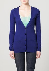 Blå stickad cardigan med en djup V-ringning, fem svarta knappar och två framfickor. Slim fit med ribbade ärmslut och nederkant.