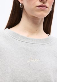 Gros plan sur une personne portant des boucles d'oreilles argentées en forme d'anneaux et un sweat-shirt gris clair avec le logo "Boss" brodé en blanc sur la poitrine.