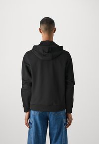 Sweatshirt à capuche noir avec une coupe décontractée, des poignets et un ourlet côtelés. La vue arrière montre une texture de tissu lisse et des lignes épurées, porté avec un jean bleu ample.