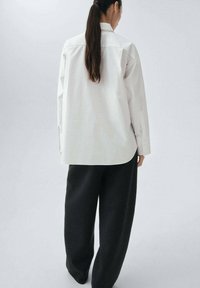 WeißesButton-up-Shirt mit einem schlanken Design und langen Ärmeln, kombiniert mit locker sitzenden, dunkelgrauen Hosen. Sichtbare glatte Stofftexturen.