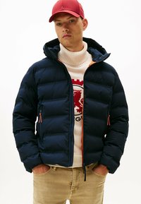 Chaqueta de plumas azul marino con capucha, con detalles en cremallera naranja y bolsillos laterales. Llevada sobre un suéter de punto crema con un diseño gráfico rojo.