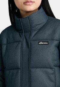 Giacca a vento blu scuro con colletto alto, caratterizzata da una superficie testurizzata e un motivo geometrico, con il logo del marchio sul petto.