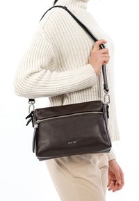 Borsa a tracolla in pelle marrone scuro con chiusura a zip, tracolla regolabile e texture liscia. Presenta un logo argentato e dettagli hardware minimi.